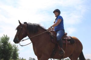 Woman in a blue top, jeans, half-chaps and riding hat riding a large bay draft cross horse in western tack
